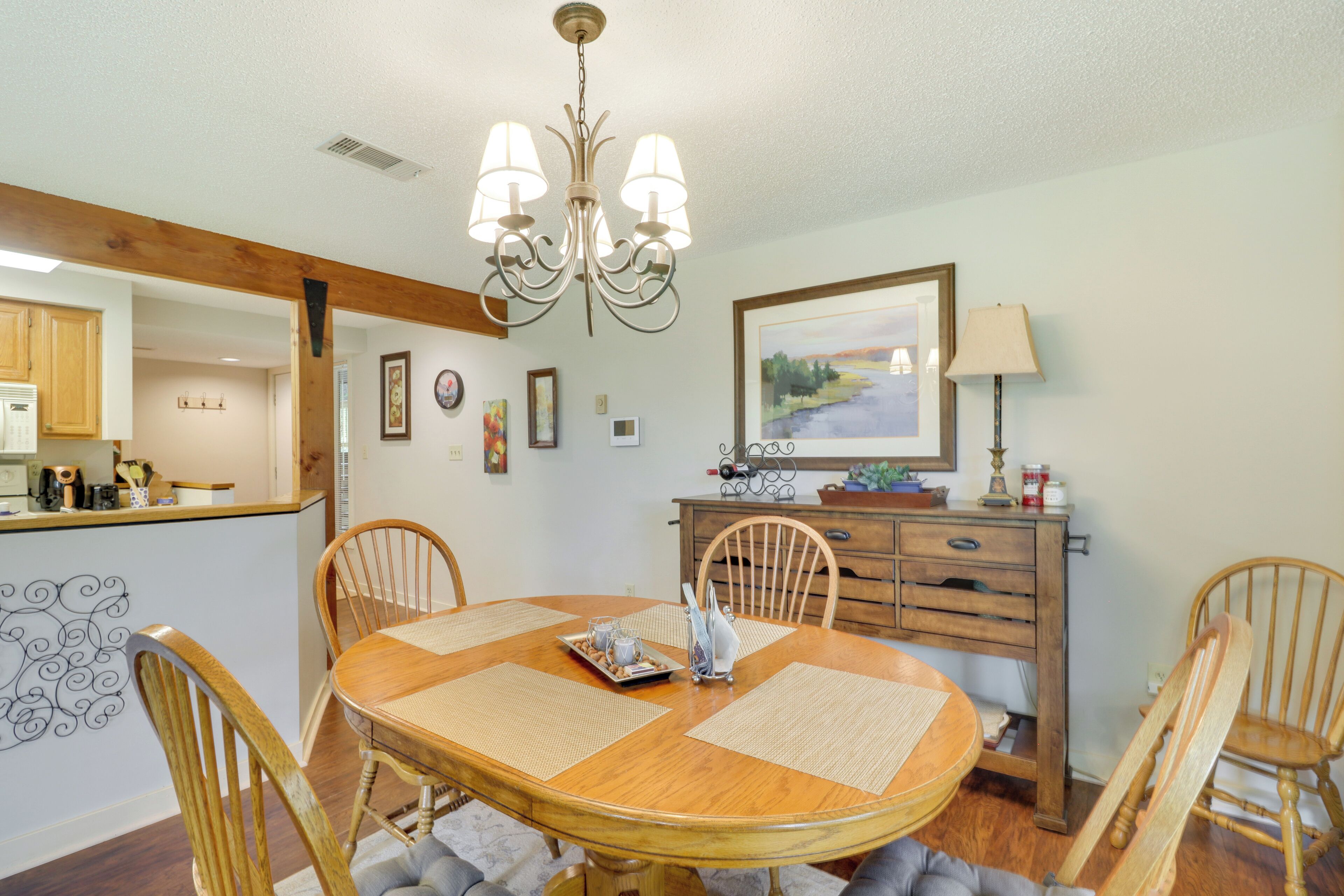 A dining area with a wooden table, chairs, and a chandelier. There is a kitchen in the background with wooden cabinets and a countertop.