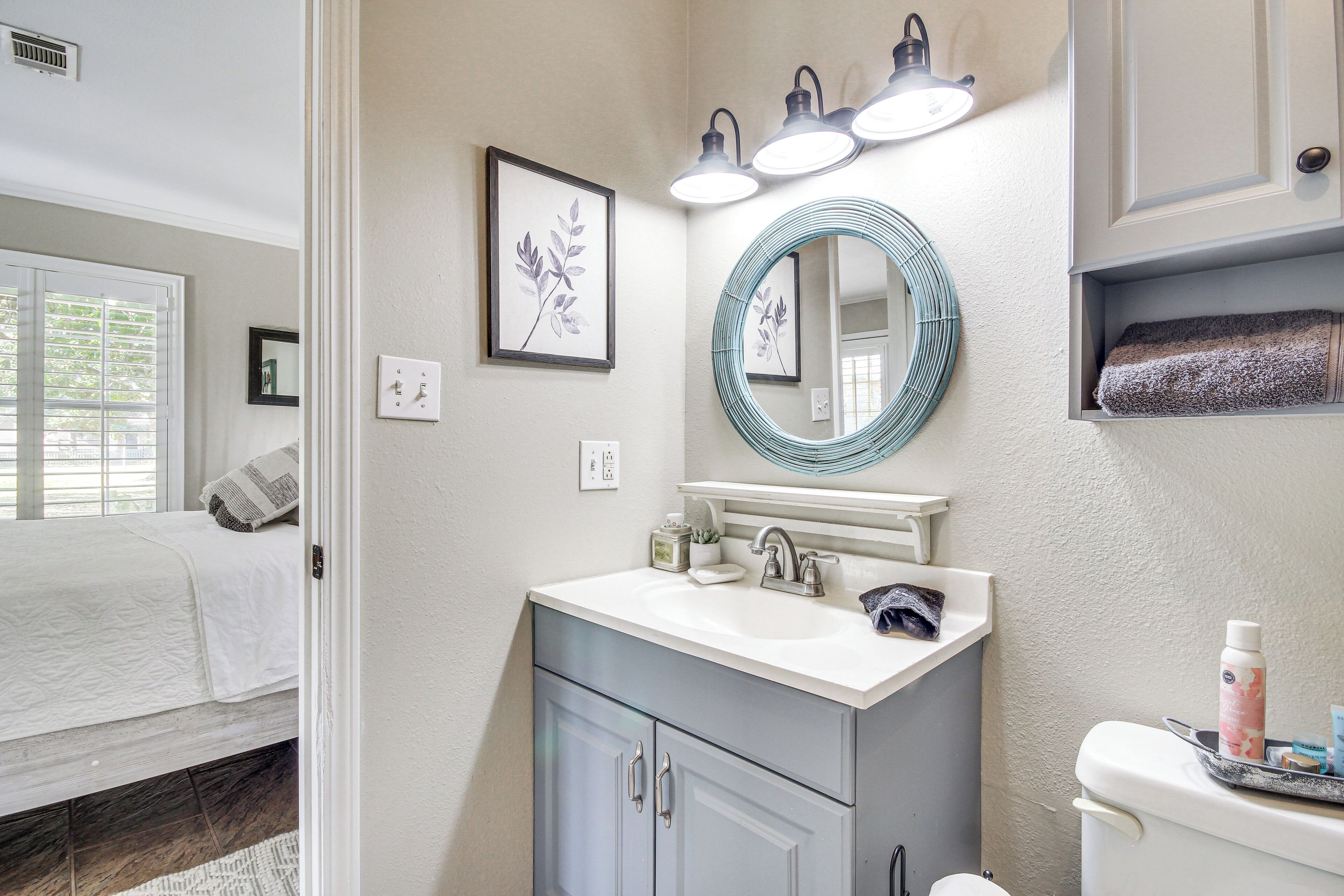 A bathroom with a sink, mirror, and toilet. There is a bed visible in the adjacent room.