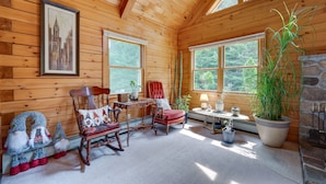 A cozy cabin interior with wooden walls, a rocking chair, a red chair, a table with a lamp, and a large window with a view of trees. - Pet-friendly Conway Cabin ~ 5 to Campton Mountain! (Thornton)