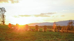 A sunset view with several wooden Adirondack chairs arranged in a semi-circle on a grassy field, with mountains in the background.