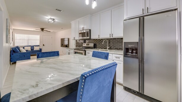 A modern kitchen with white cabinets, a stainless steel refrigerator, a microwave, and a dining area with a marble table and blue chairs. - Modern Home in Edinburg: Half-mi Walk to Utrgv! (Edinburg)