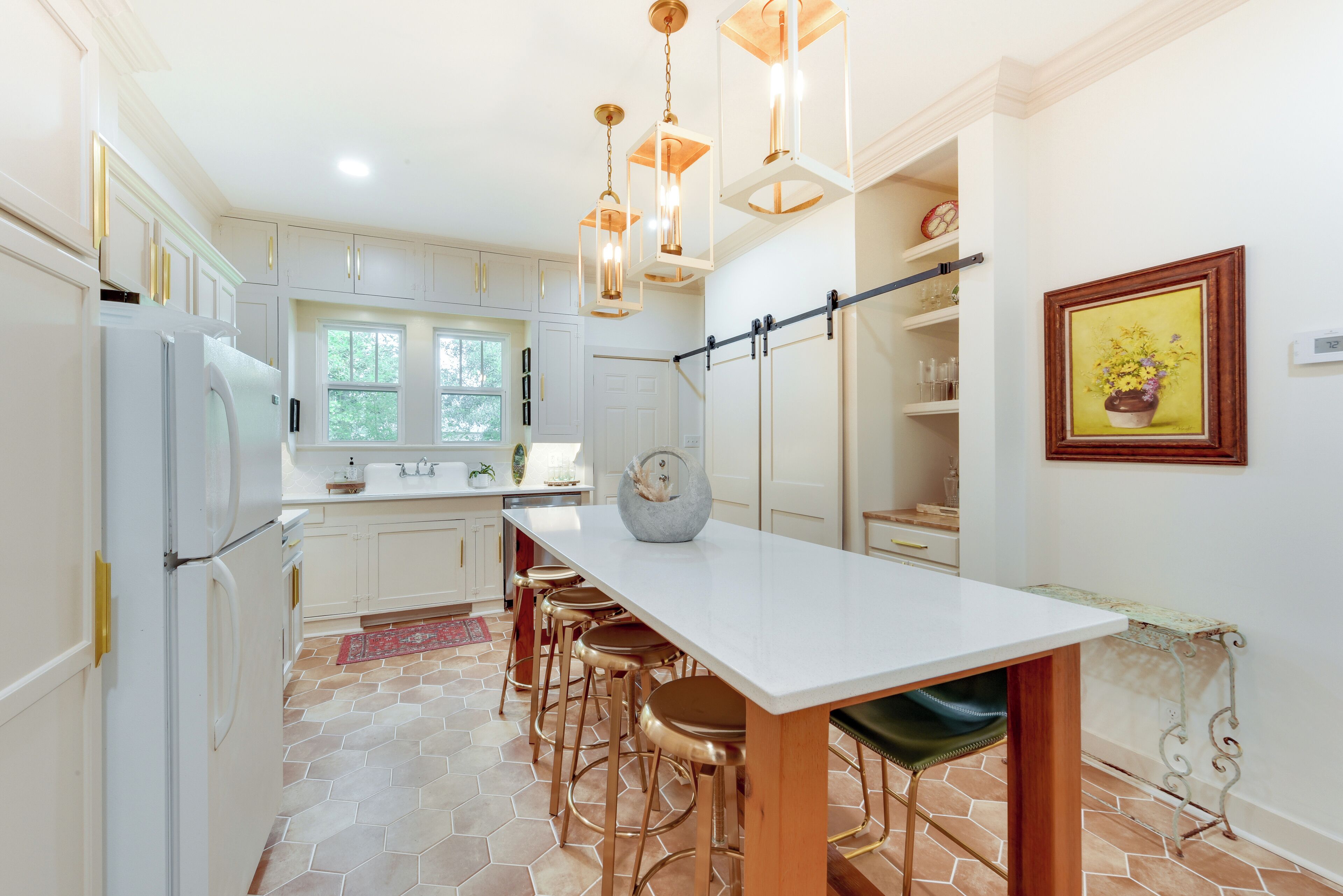 A modern kitchen with white cabinets, a large island with stools, pendant lighting, and a painting on the wall.