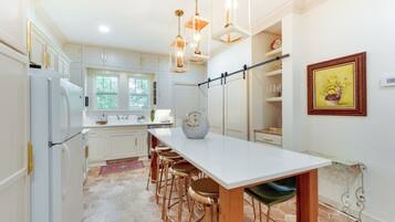 A modern kitchen with white cabinets, a large island with stools, pendant lighting, and a painting on the wall.