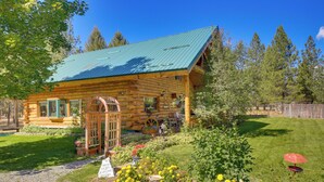 Eine Blockhütte mit einem grünen Metalldach, umgeben von einem gepflegten Garten mit verschiedenen Pflanzen und Blumen.