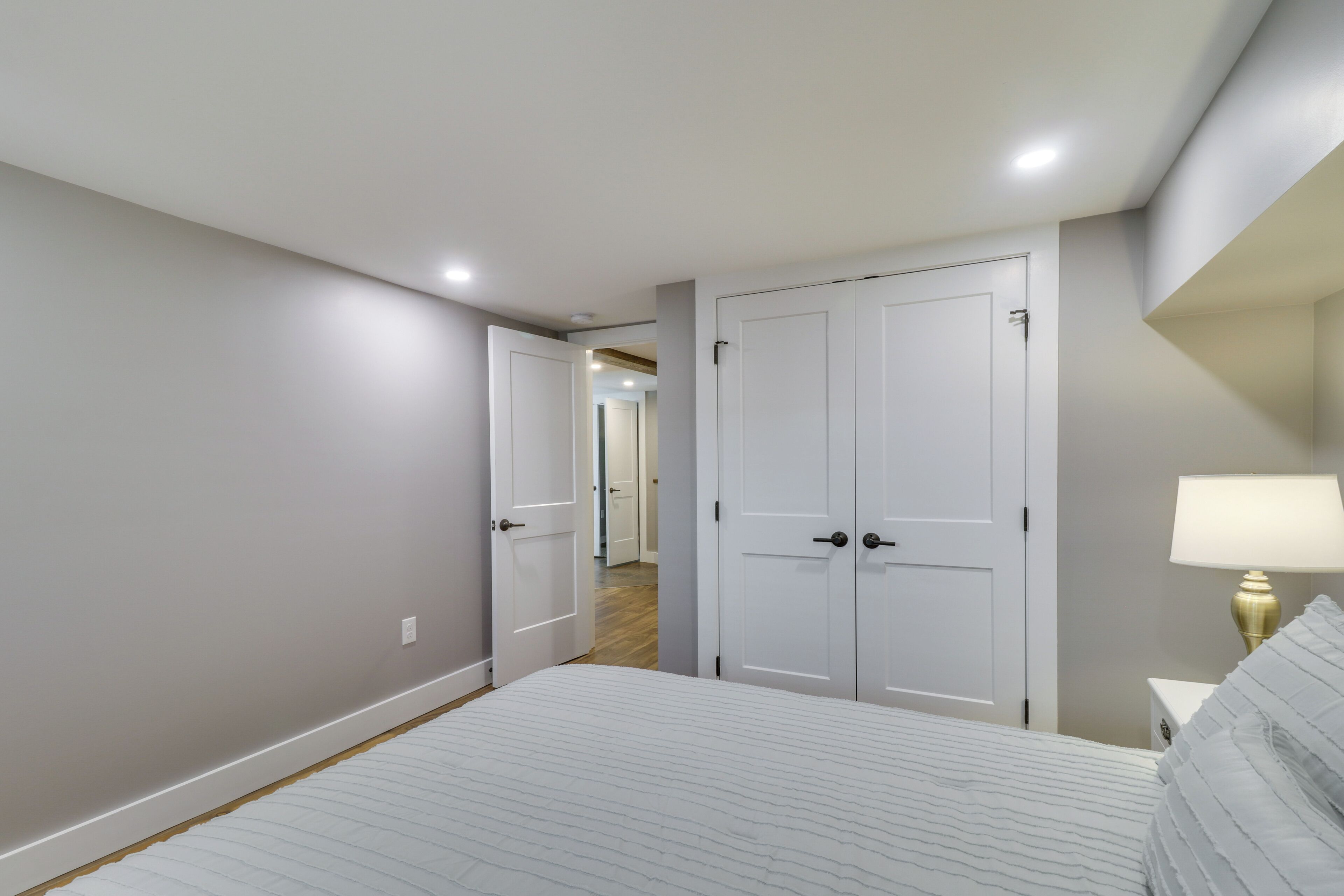 A bedroom with a bed, two white doors, a nightstand with a lamp, and a hallway leading to another room.