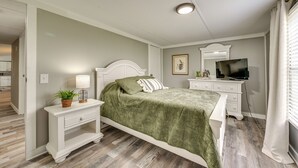 Un dormitorio con cama, mesitas de noche, cómoda, televisor y ventana con cortinas.