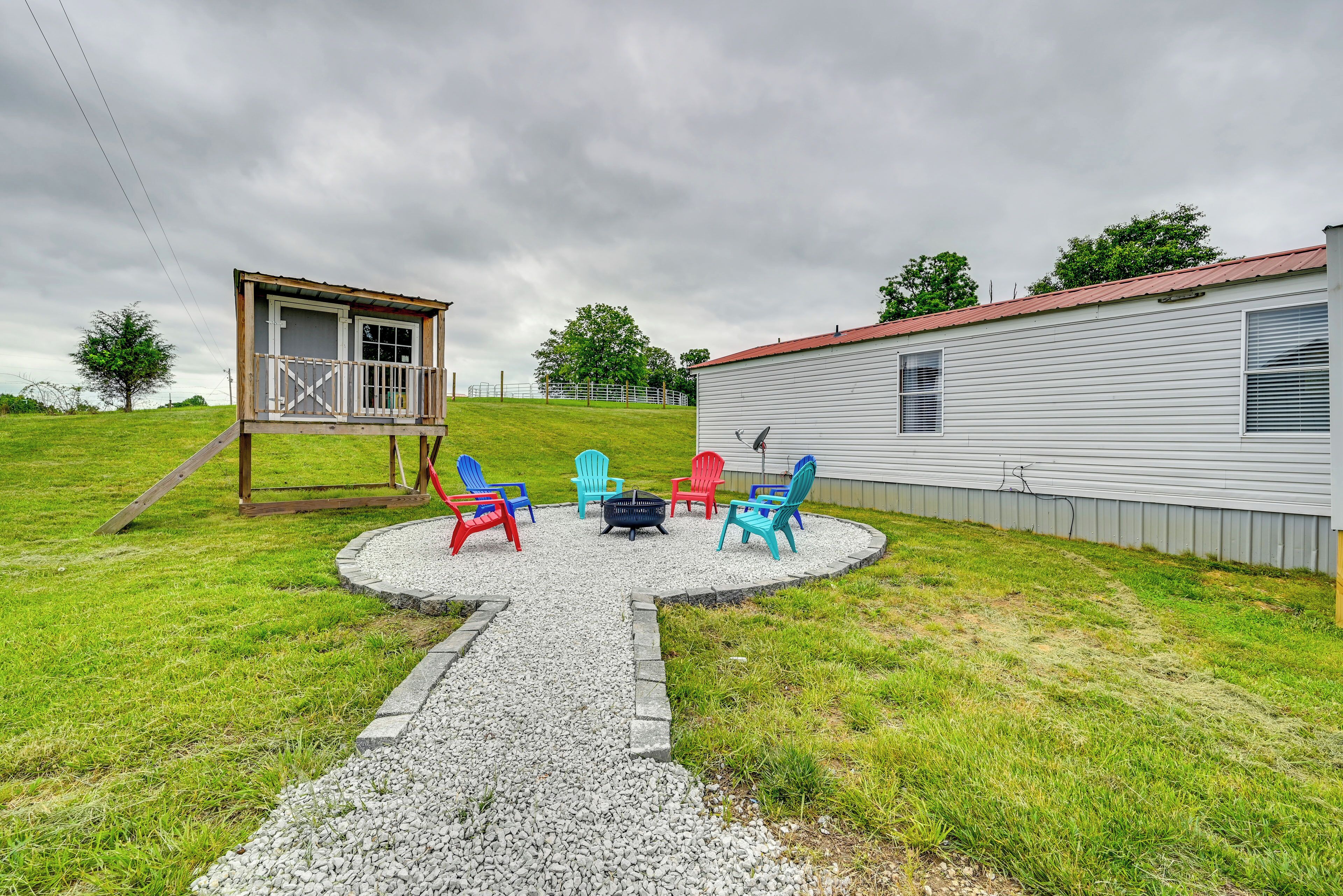 A backyard with a gravel patio, colorful Adirondack chairs, a fire pit, and a small wooden structure.