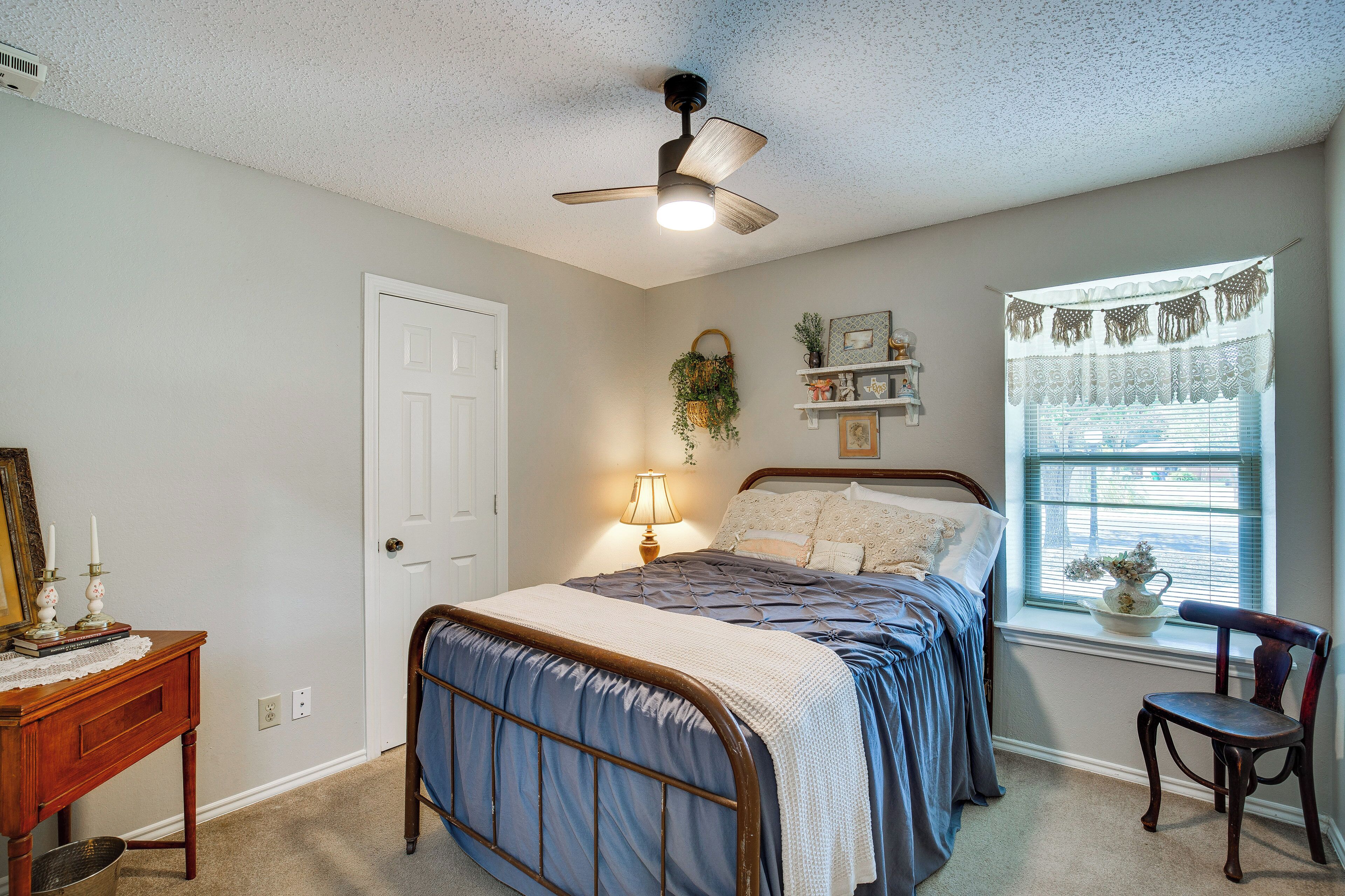 A bedroom with a bed, a desk, a chair, a ceiling fan, a window with curtains, and a wall-mounted shelf with decorative items.