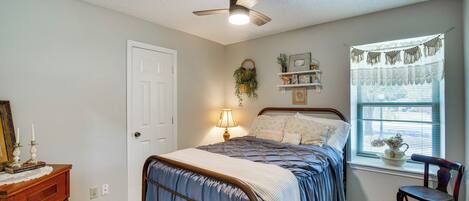 A bedroom with a bed, a desk, a chair, a ceiling fan, a window with curtains, and a wall-mounted shelf with decorative items.