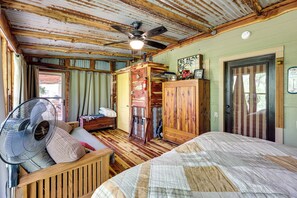 A rustic room with a wooden ceiling, a bed with a quilt, a wooden bench, a fan, and a door with a striped curtain. - Flag Hole riverfront Cabin Near Local Attractions! (Caddo Gap)