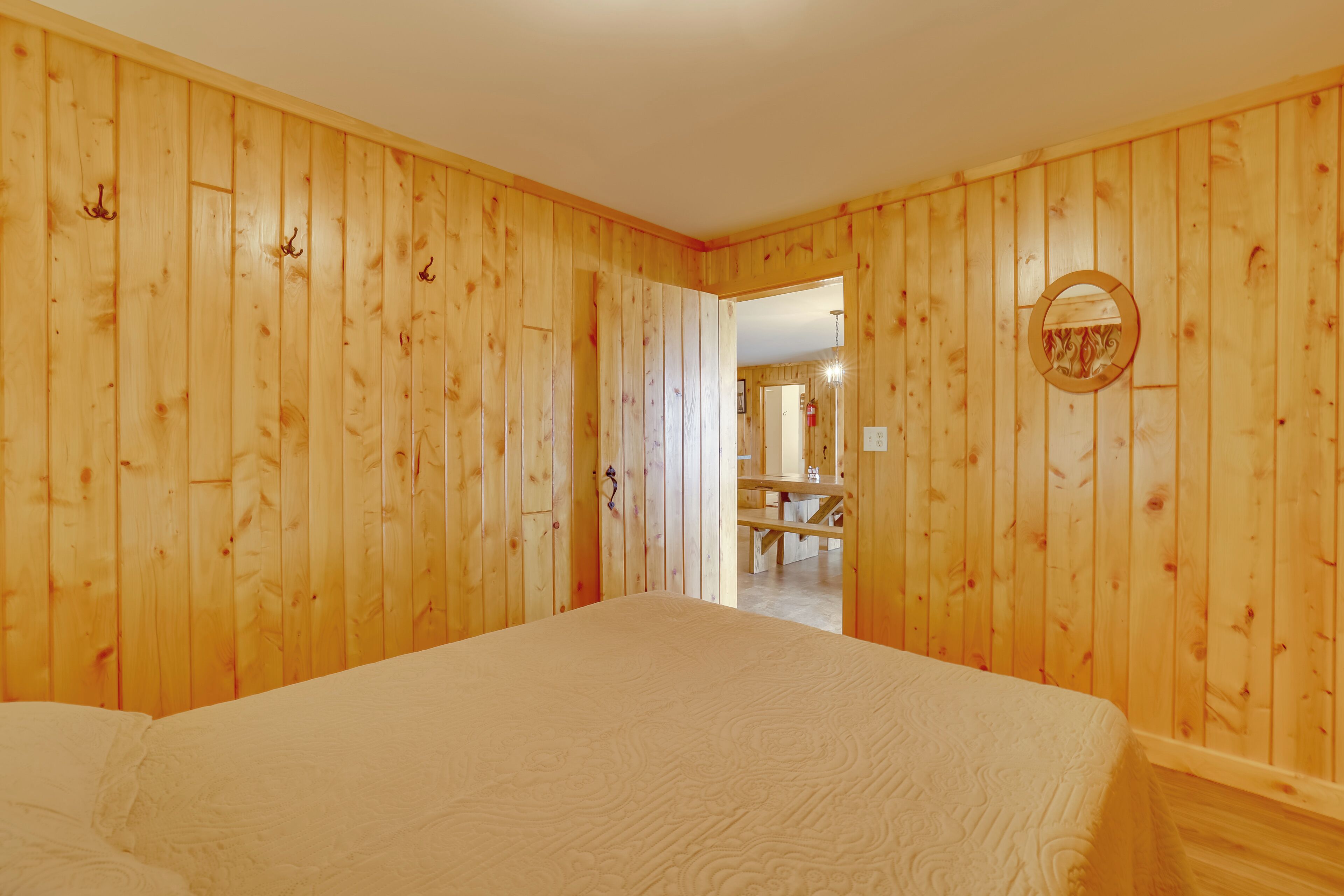 A room with wooden panel walls, a bed with a white bedspread, a round mirror on the wall, and a doorway leading to a dining area.