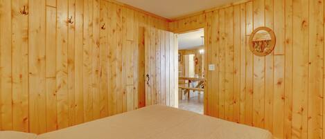 A room with wooden panel walls, a bed with a white bedspread, a round mirror on the wall, and a doorway leading to a dining area.