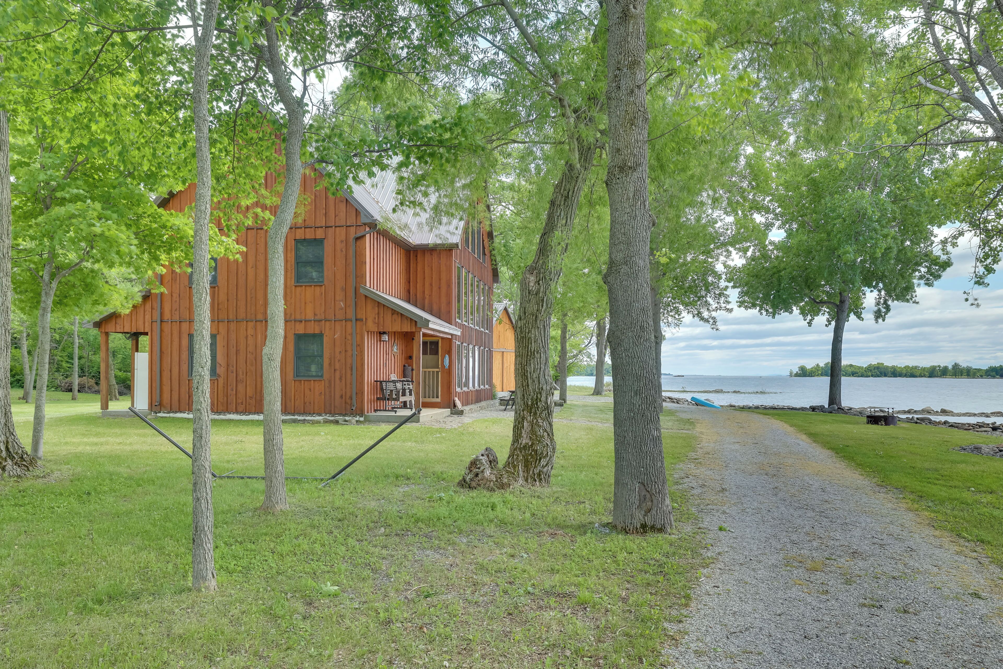 A wooden cabin with a porch, surrounded by trees and a gravel path leading to a lake.