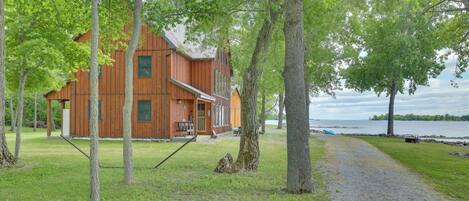 A wooden cabin with a porch, surrounded by trees and a gravel path leading to a lake.