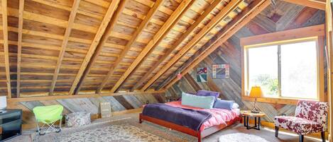 A cozy attic bedroom with a wooden ceiling, a bed with colorful bedding, a chair, and a rug.