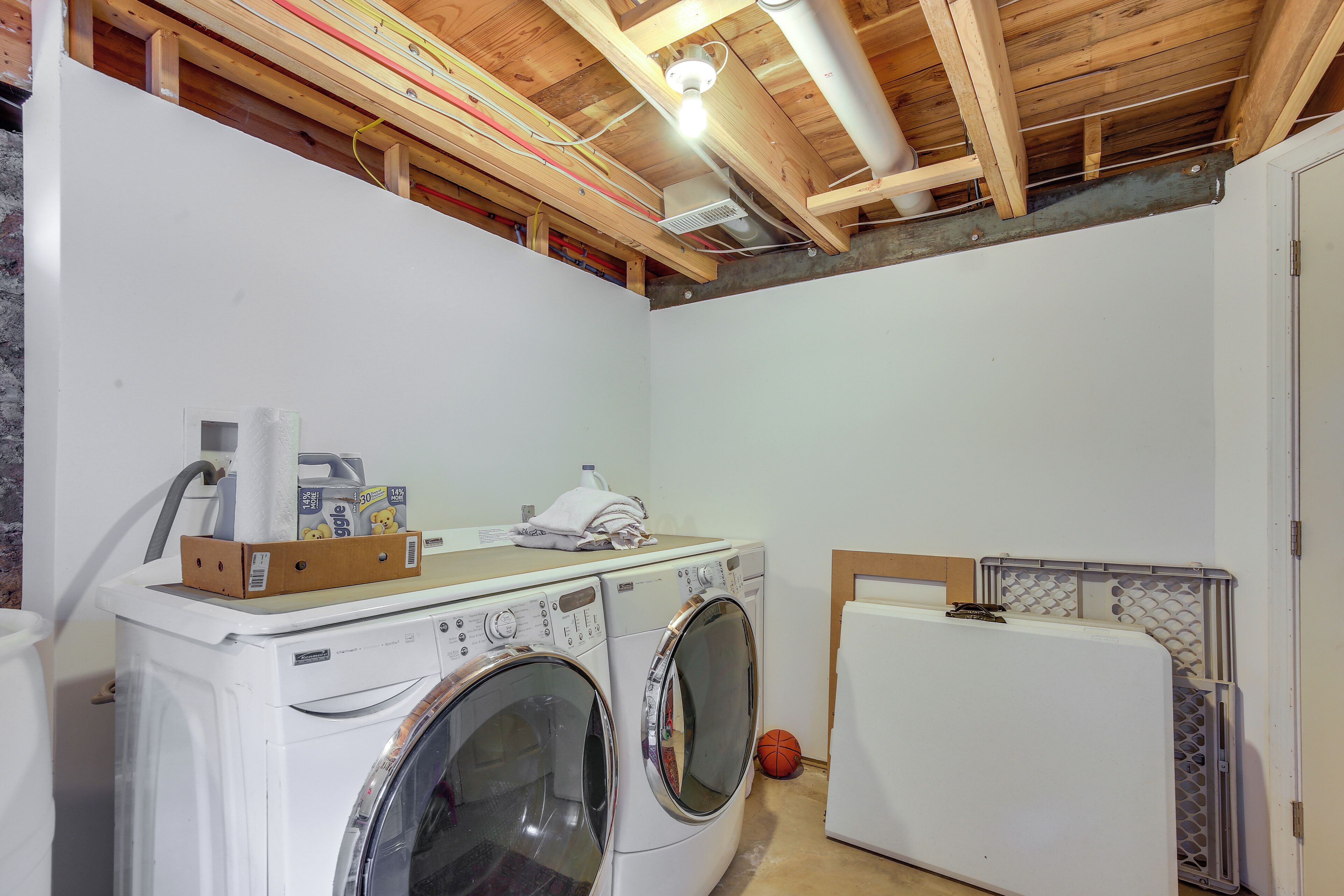 A laundry room with two white front-loading washing machines, a dryer, and a wooden ceiling with exposed beams.