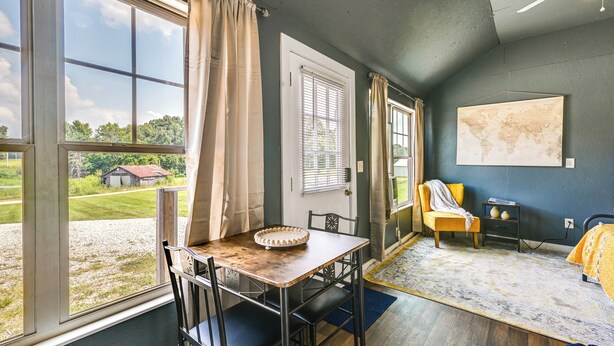 Une pièce avec une grande fenêtre, une table à manger avec des chaises, un lit recouvert d’une couverture jaune, un fauteuil avec un coussin, une petite table sur laquelle repose un globe terrestre, et une carte accrochée au mur.