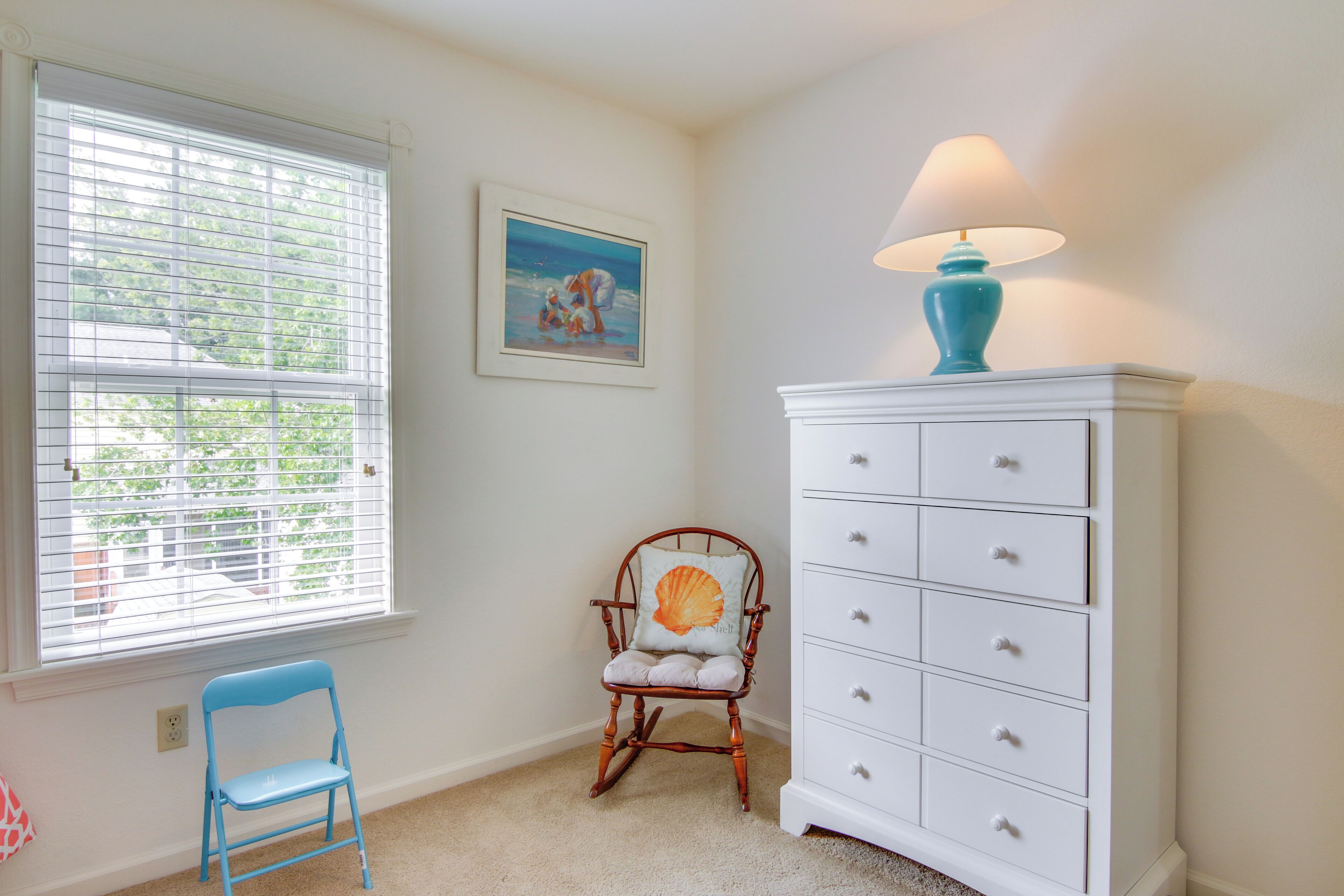 A room with a rocking chair, a white dresser with drawers, a lamp, and a window with blinds.