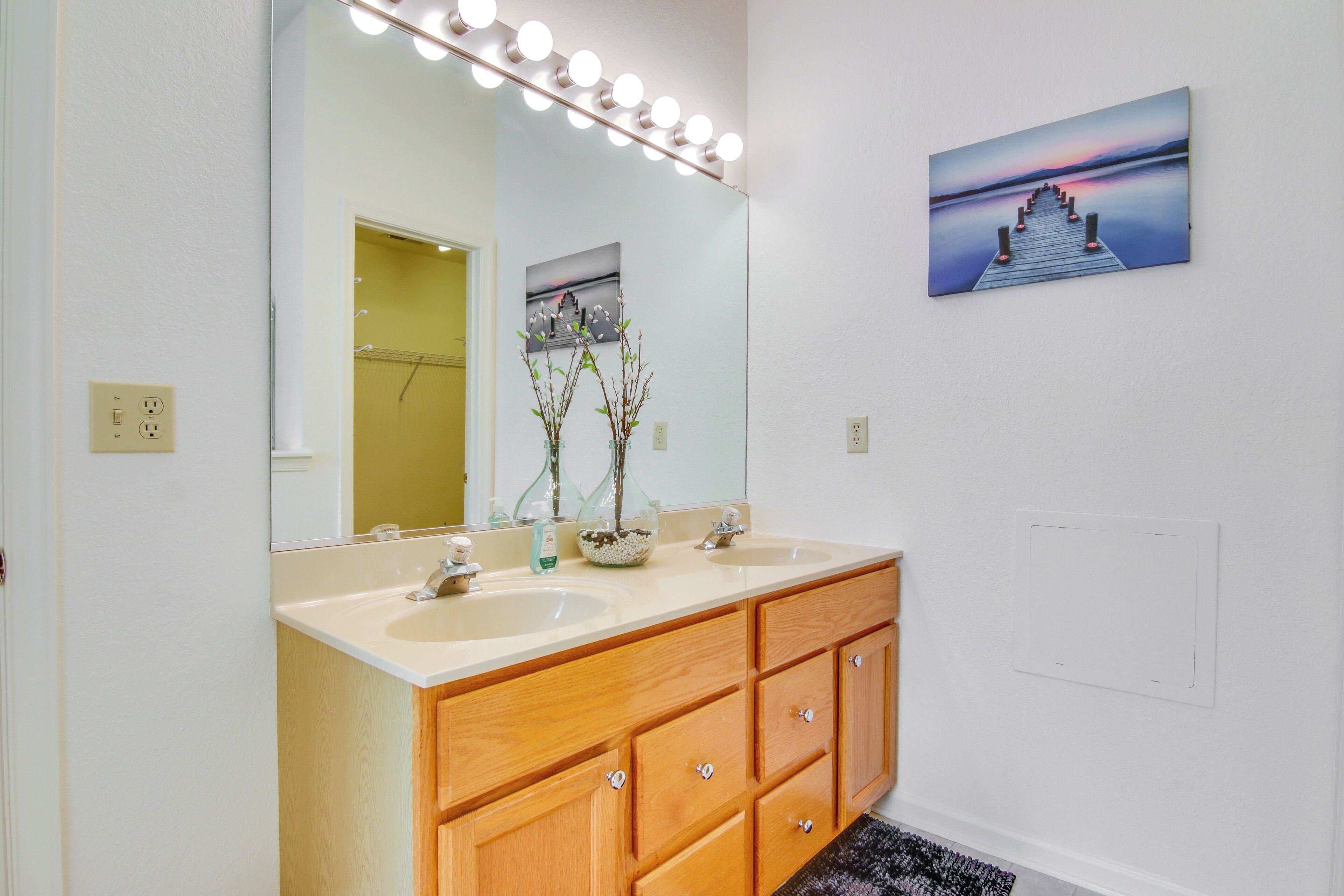 A bathroom with two sinks, a large mirror with lights, wooden cabinets, and a framed picture on the wall.
