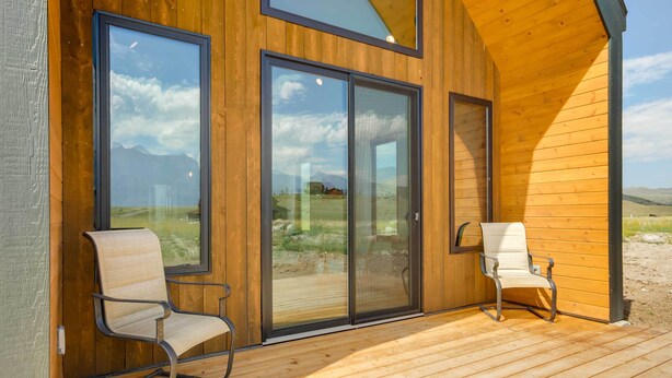 A wooden cabin with large sliding glass doors, two chairs on a deck, and a view of mountains. - Yellowstone Prairie Retreat: 360 Mountain Views! (Emigrant)