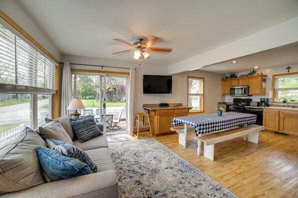 A spacious living area with a sofa, a coffee table, and a dining table. There is a kitchen with wooden cabinets and a microwave. A ceiling fan is visible, and a television is mounted on the wall. - Walk to Beach, Lake Edinboro Views: 'camp Dickson' (Edinboro)
