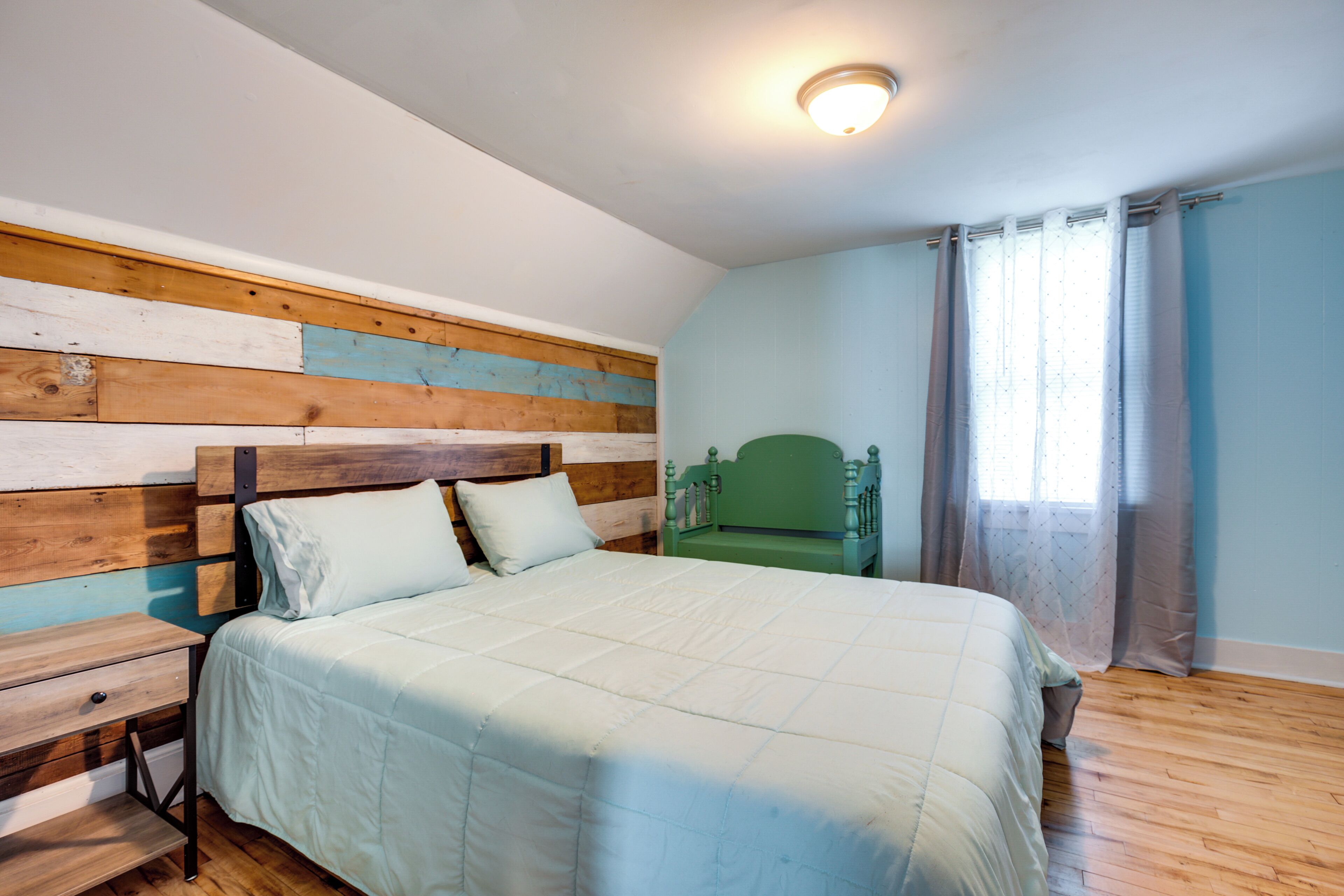 Una camera da letto con una testiera in legno, un letto con copripiumino bianco, un comodino e una finestra con tende.
