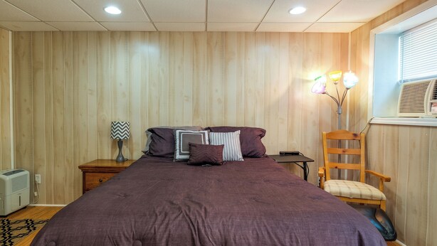 A bedroom with a large bed, wooden walls, a wooden chair, and a bedside table with a lamp. - Cozy Hiwassee Vacation Rental w/ Yard (Hiwassee)