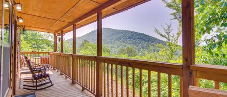 A wooden deck with a railing, rocking chairs, and a view of a forested mountain.
