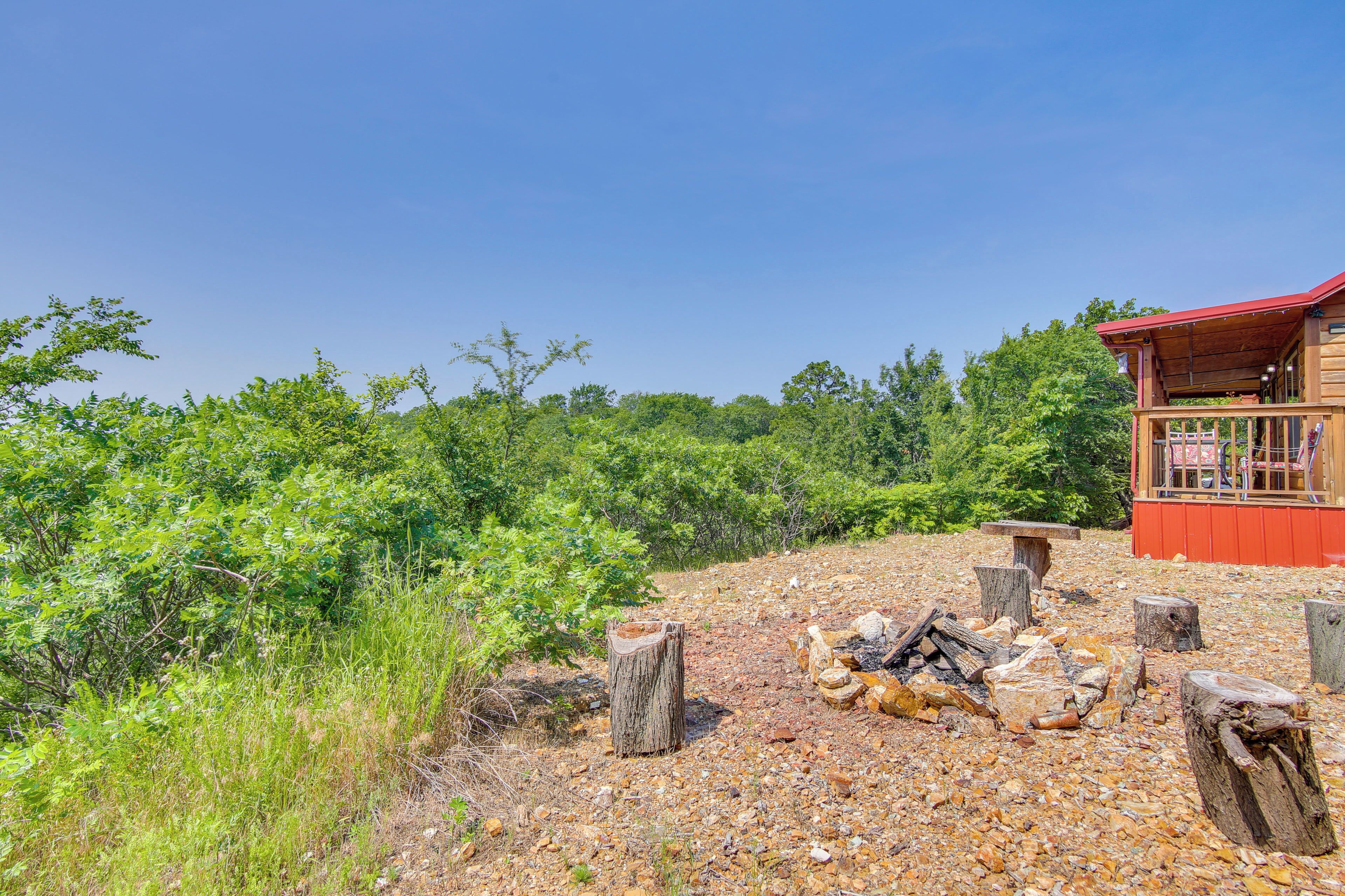A rustic cabin with a red roof, surrounded by dense greenery and a gravel path leading to a fire pit area.