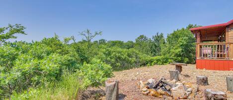 A rustic cabin with a red roof, surrounded by dense greenery and a gravel path leading to a fire pit area.