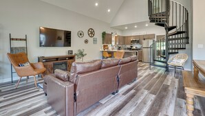 Ein geräumiges Wohnzimmer mit einem braunen Ledersofa, einem Couchtisch aus Holz und einer hohen Decke mit Einbaustrahlern. Im Hintergrund ist eine Küche mit modernen Geräten und einer Wendeltreppe zu sehen.