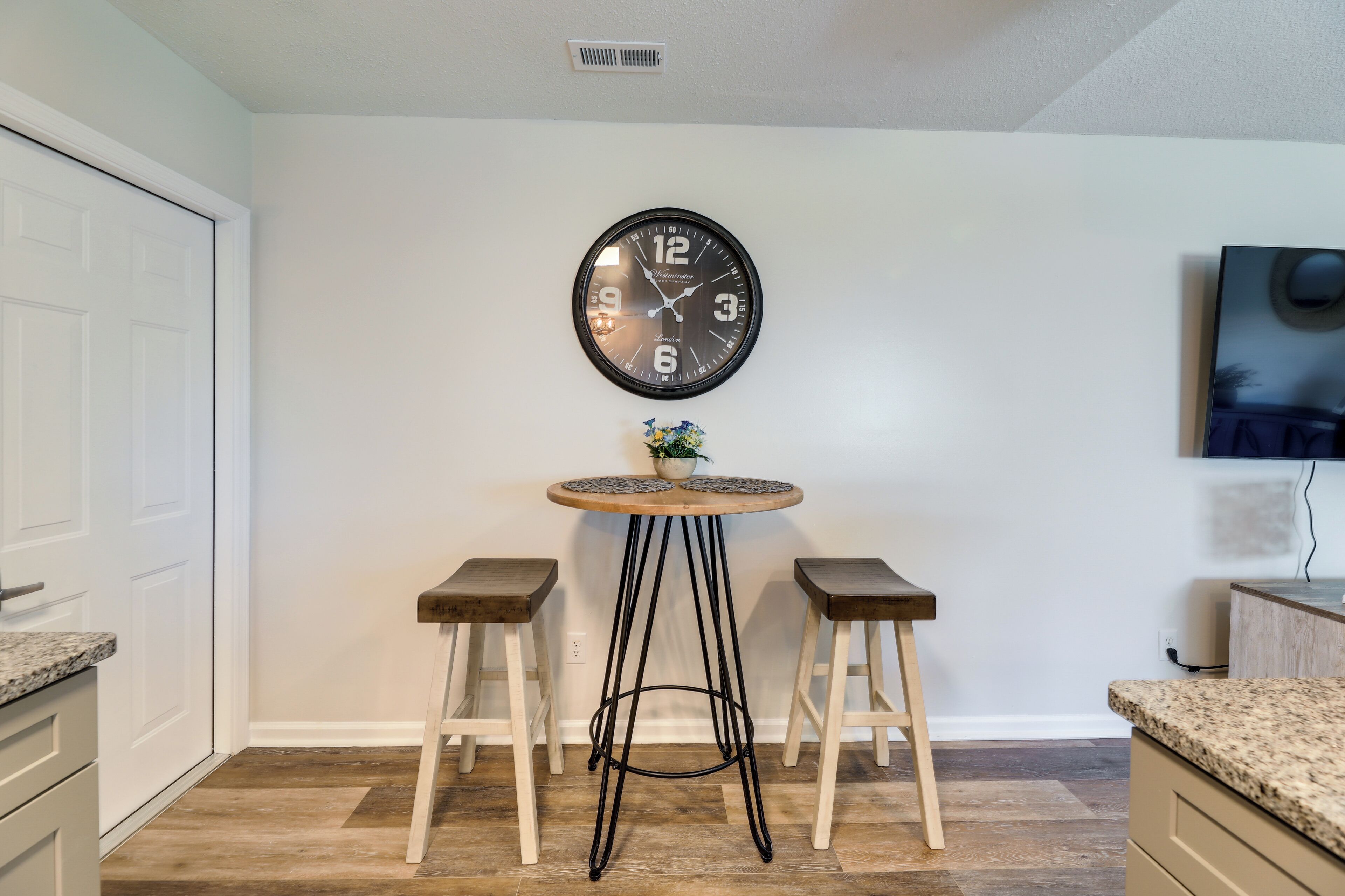 A kitchen with a round wooden table, two bar stools, a wall-mounted clock, and a small potted plant.
