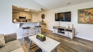 Cocina moderna con microondas, fregadero y taburetes de bar. Hay una mesa de comedor con un centro de mesa con planta y un televisor montado en la pared.