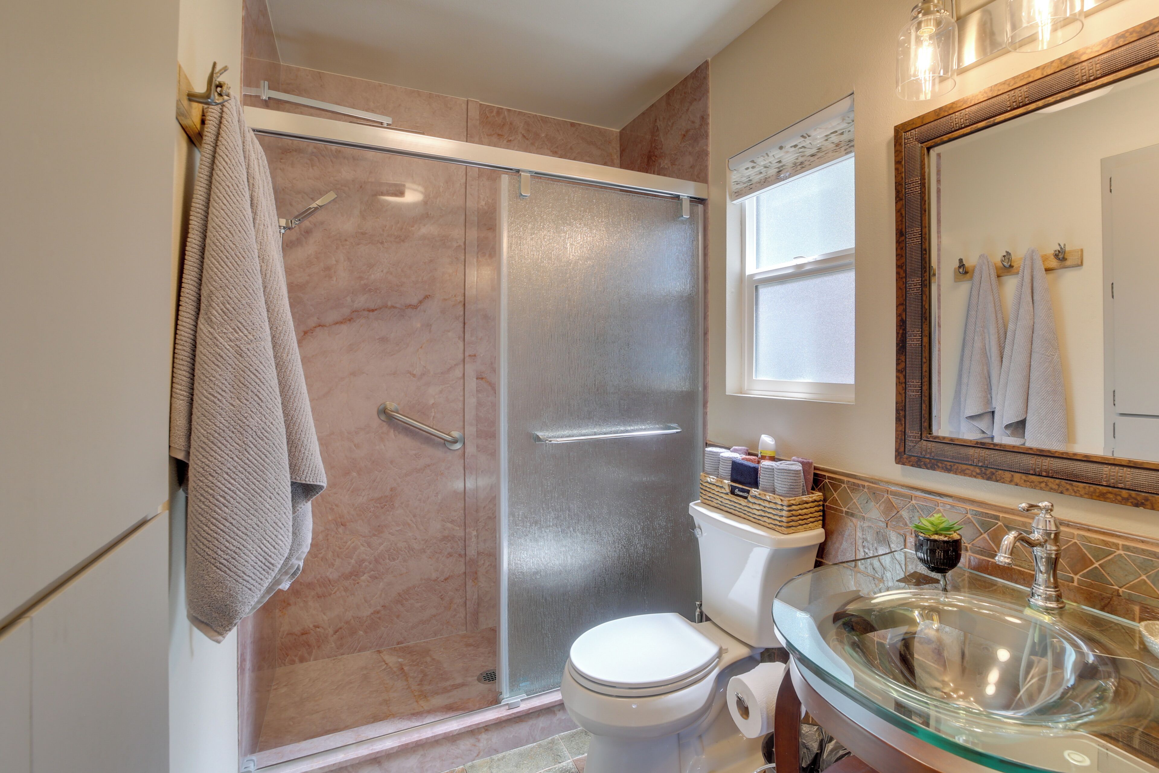 A bathroom with a glass shower enclosure, a toilet, a glass sink, and a mirror with towels hanging on the wall.