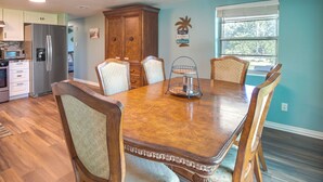 A dining area with a wooden table and chairs, a kitchen with a refrigerator and cabinets, and a window with blinds. - Cookson Vacation Rental w/ Spacious Yard & Porch! (Cookson)