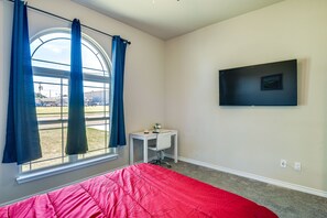A bedroom with a large window, a bed with a red blanket, a small desk with a chair, and a flat-screen TV mounted on the wall.