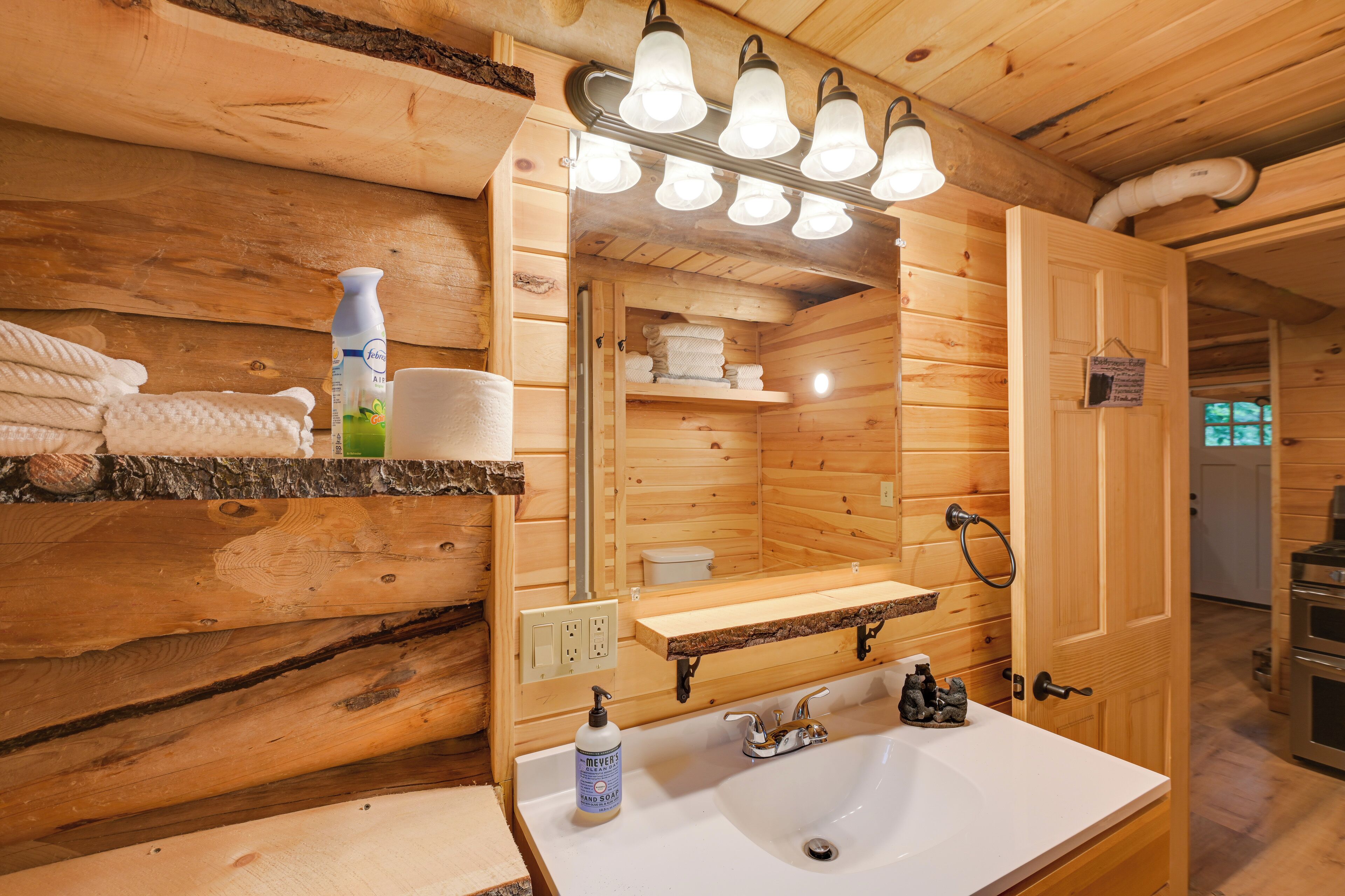 Une salle de bain rustique, aménagée dans une cabane en bois, avec un lavabo, un miroir et des étagères où sont rangées les serviettes et les articles de toilette.