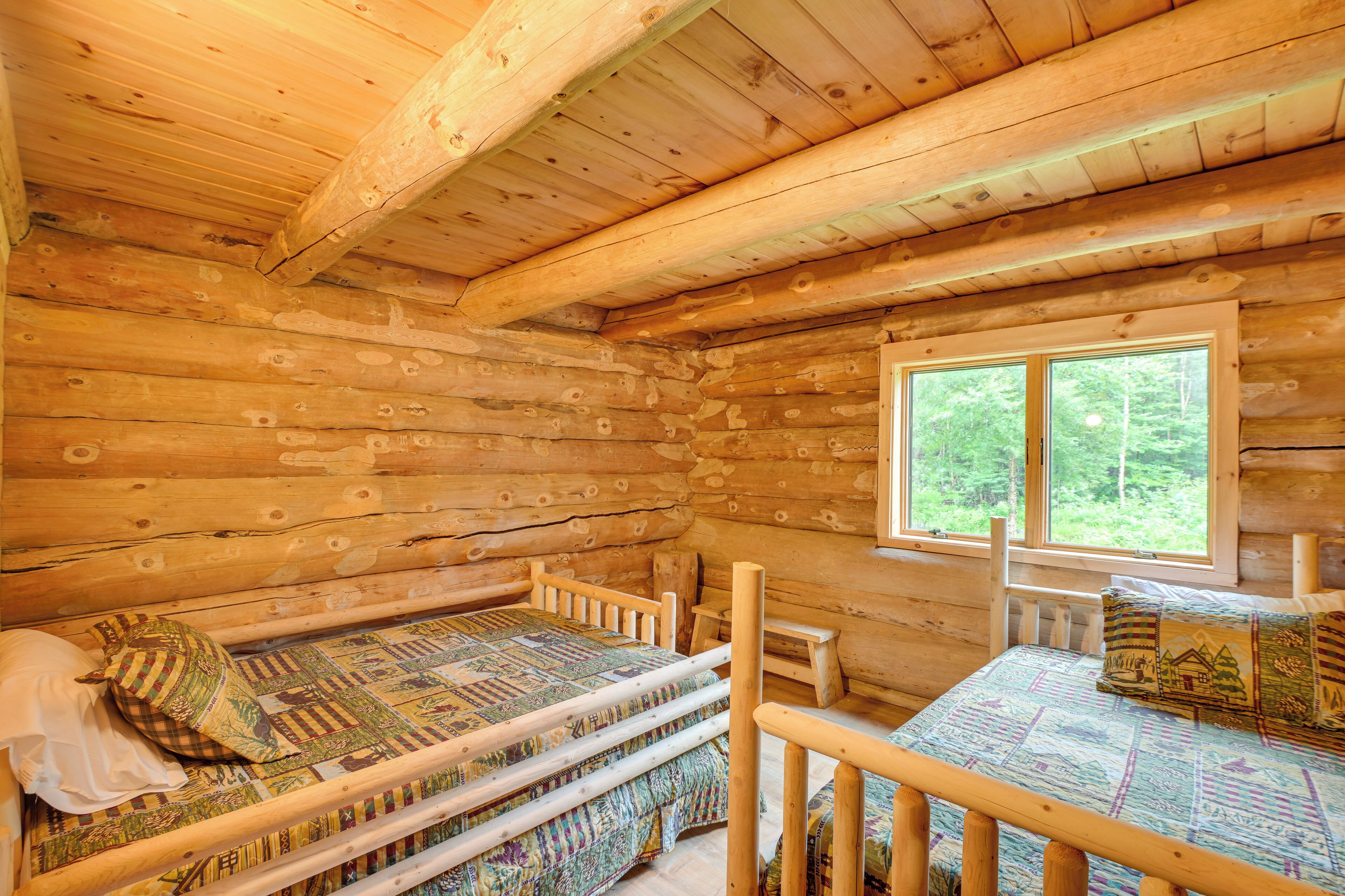 A log cabin room with two wooden beds, a window with a view of the forest, and a wooden ceiling.