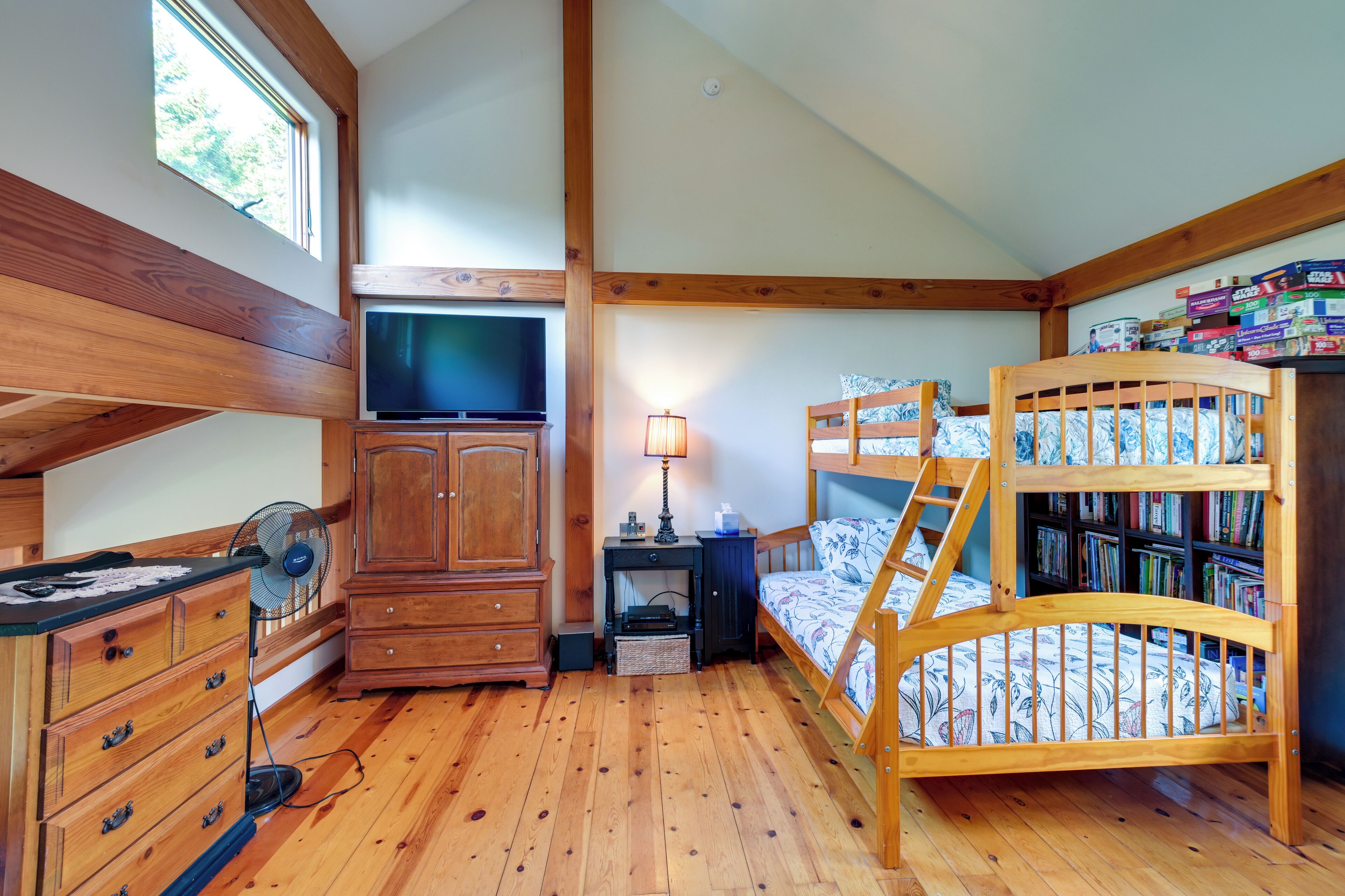 Una habitación con piso de madera, una cama nido, un televisor, un escritorio con lámpara y una estantería.