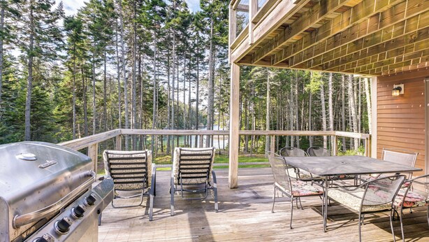 Una terraza de madera con una parrilla, sillas y una mesa, con vistas a un área boscosa.