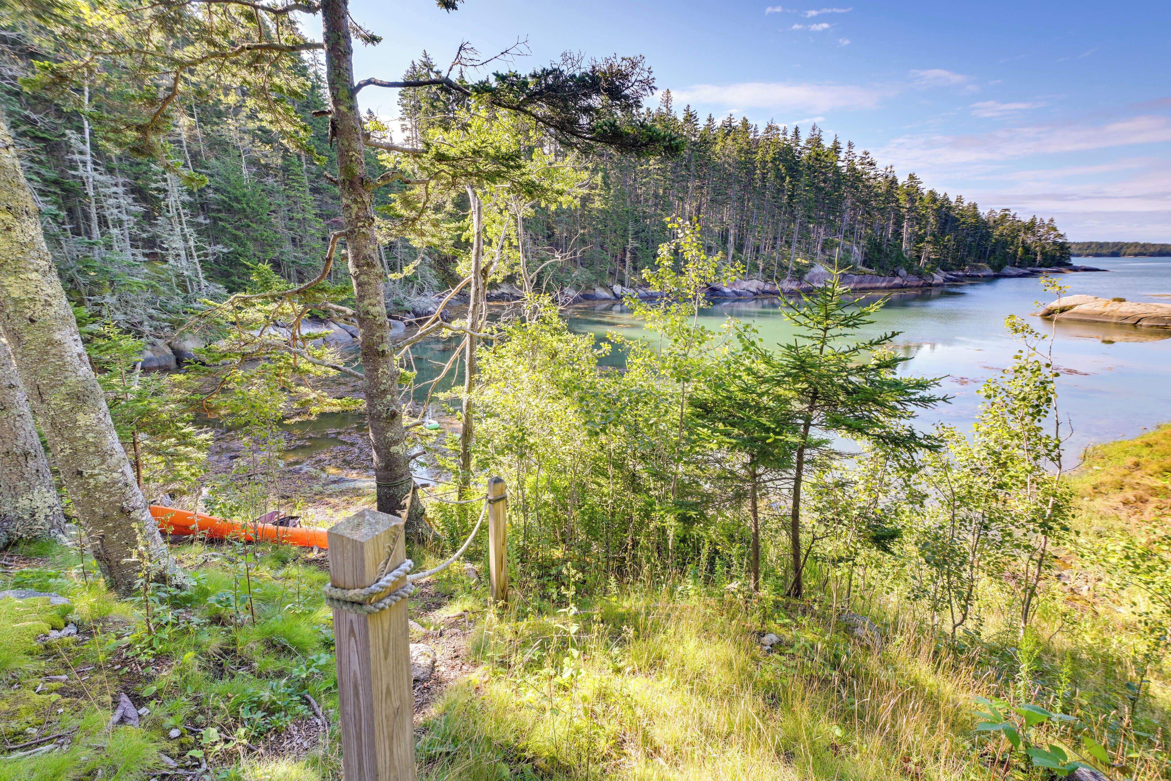 A forested area with a wooden post and rope barrier, a canoe, and a calm body of water.