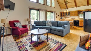 A spacious living room with a gray sectional sofa, a red armchair, and a round coffee table. A kitchen with wooden cabinets and a black refrigerator is visible in the background. - Cabin w/ Deck ~ 3 Mi to Apple Valley Lake! (Howard)
