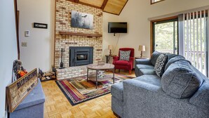 A cozy living room with a brick fireplace, a comfortable sofa, a red armchair, and a television mounted on the wall. - Cabin w/ Deck ~ 3 Mi to Apple Valley Lake! (Howard)