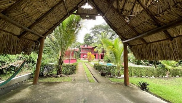 Une allée couverte mène à un bâtiment rouge, entouré d’une végétation luxuriante.