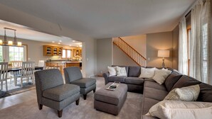 Un salon spacieux avec un canapé gris, deux fauteuils et une table basse. Un escalier mène à un niveau supérieur, et on aperçoit un coin repas au fond de la pièce.