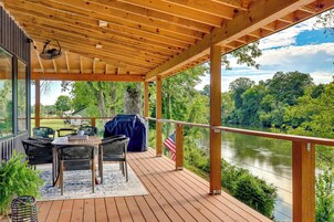 Terraza de madera con mobiliario exterior, una bandera y vistas al río.