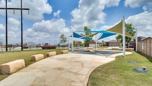 Uma trilha pavimentada leva a uma área de estar coberta por um toldo azul, cercada por uma grade e edifícios residenciais ao fundo.