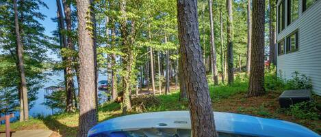 Un kayak est attaché à un arbre dans une zone boisée, avec un lac et un chalet visibles au loin.