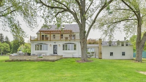 A two-story house with a balcony, a large tree in the front yard, and a well-maintained lawn. - Pistakee Lake Estate w/ Boat Dock & Hot Tub! (McHenry)