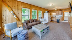 A cozy cabin with a living area featuring a sofa, a coffee table, and a patterned ottoman. A ski lift is mounted on the wall, and the kitchen area is visible in the background. - Lakefront Grant Cottage w/ Grill & Fire Pit! (Grant)